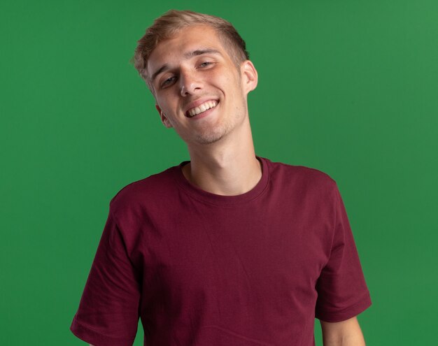 Sonriendo inclinando la cabeza joven guapo con camisa roja aislado en la pared verde