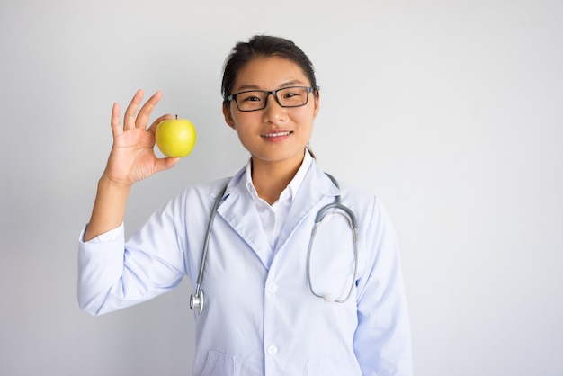 Sonriendo el joven médico femenino asiático que muestra la manzana. Concepto de nutrición saludable.