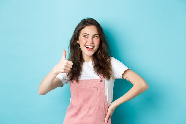 Sonriendo a una linda chica adolescente con un hermoso maquillaje de rubor y glamour, mostrando el pulgar hacia arriba en señal de aprobación, mire el banner de la esquina superior izquierda, recomendando la tienda, de pie contra el fondo azul.