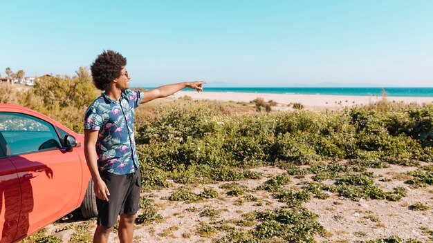 Sonriendo masculino étnico joven permanente por la orilla del mar y mostrando la dirección