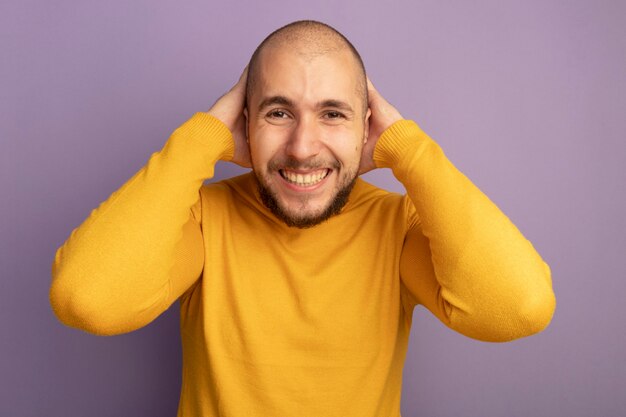 Sonriendo mirando hacia adelante chico guapo joven poniendo las manos detrás de la cabeza aislada en púrpura