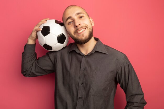 Sonriendo mirando hacia adelante chico guapo joven poniendo la pelota en el hombro aislado en rosa