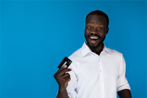 Sonriendo mirando hacia adelante hombre afroamericano en camisa blanca tiene tarjeta de crédito en una mano