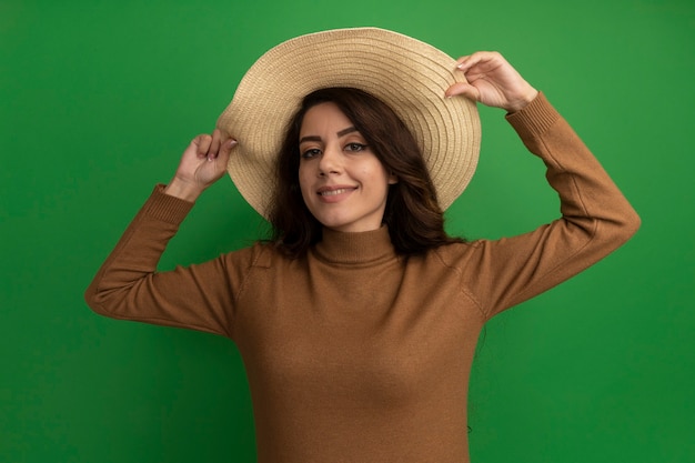 Sonriendo mirando al frente hermosa joven vistiendo y sosteniendo el sombrero aislado en la pared verde