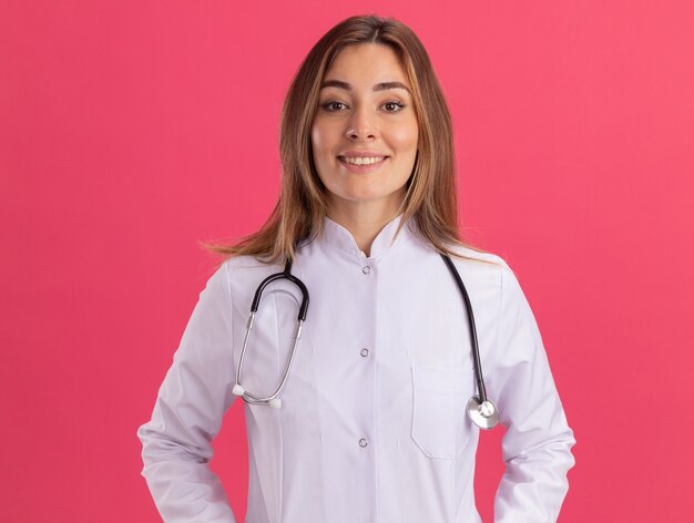 Sonriendo mirando al frente joven doctora vistiendo bata médica con estetoscopio aislado en la pared rosa