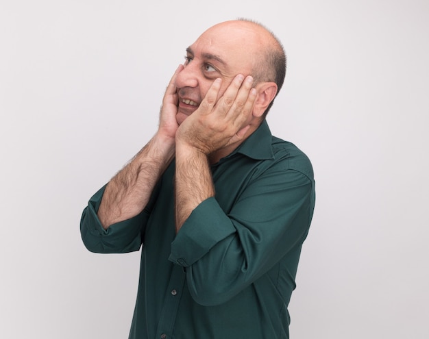 Sonriendo mirando al lado hombre de mediana edad vestido con camiseta verde poniendo las manos en las mejillas aisladas en la pared blanca