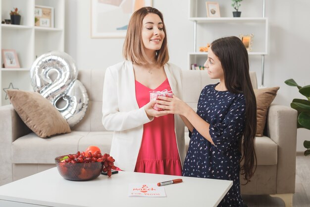 Sonriendo mirando el uno al otro hija da presente a la madre en el día de la mujer feliz en la sala de estar