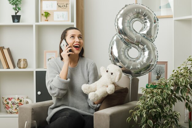 Sonriendo mirando hacia arriba hermosa niña en el día de la mujer feliz con osito de peluche habla por teléfono sentado en un sillón en la sala de estar