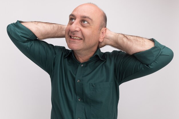 Sonriendo mirando hacia arriba el hombre de mediana edad vestido con camiseta verde poniendo la mano detrás de la cabeza aislada en la pared blanca