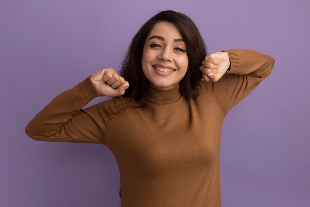 Sonriendo mirando a la cámara hermosa joven vistiendo suéter de cuello alto marrón sosteniendo puños aislados en la pared púrpura