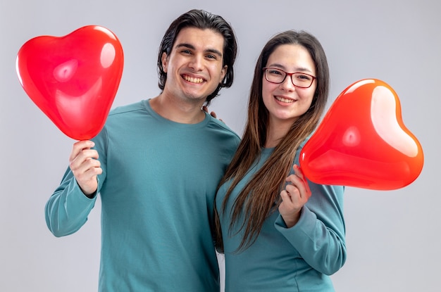 Sonriendo mirando a la cámara pareja joven en el día de San Valentín sosteniendo globos de corazón aislado sobre fondo blanco.