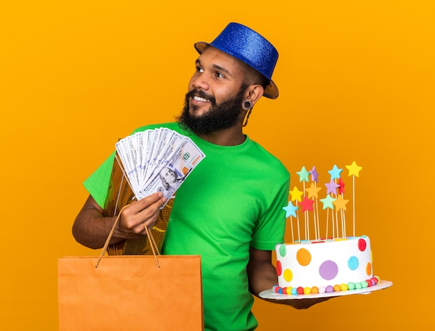 Sonriendo mirando de lado joven afroamericano vistiendo gorro de fiesta sosteniendo regalos y pastel con dinero en efectivo