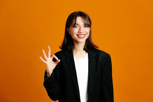 Sonriendo mostrando bien gesto joven hermosa mujer vistiendo chaqueta negra aislado sobre fondo naranja