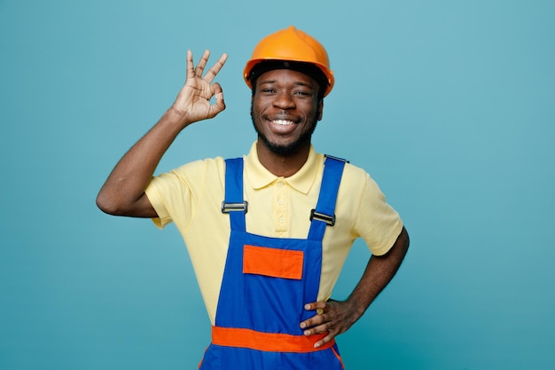 Sonriendo mostrando bien gesto poniendo la mano en las caderas joven constructor afroamericano en uniforme aislado sobre fondo azul.