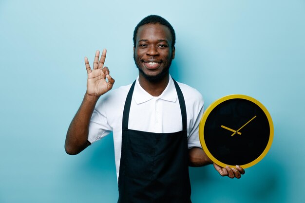 Sonriendo mostrando gesto bien sosteniendo reloj de pared joven peluquero afroamericano en uniforme aislado sobre fondo azul.