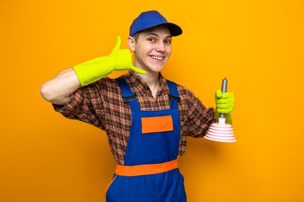Sonriendo mostrando gesto de llamada telefónica joven chico de limpieza con uniforme y gorra con guantes sosteniendo el émbolo