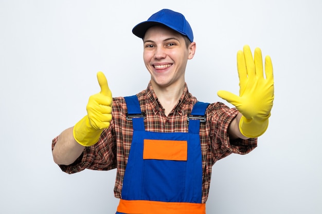 Sonriendo mostrando gesto de parada con el pulgar hacia arriba joven chico de limpieza con uniforme y gorra con guantes