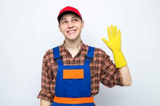 Sonriendo mostrando hola gesto joven chico de limpieza con uniforme y gorra con guantes