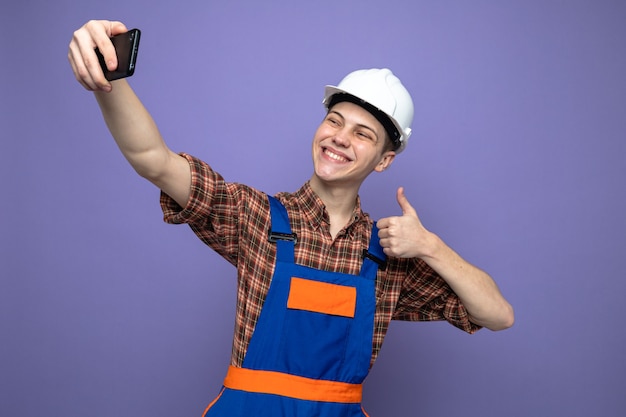 Sonriendo mostrando el pulgar hacia arriba joven constructor con uniforme tomar un selfie