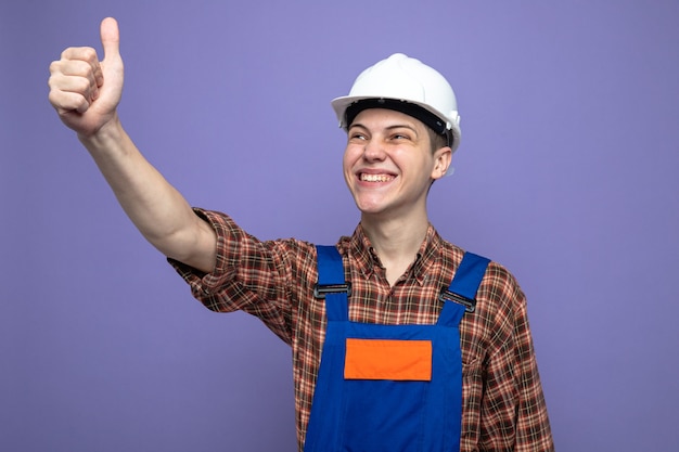 Sonriendo mostrando el pulgar hacia arriba joven constructor con uniforme