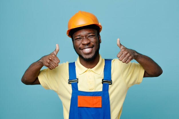 Sonriendo mostrando los pulgares hacia arriba joven constructor afroamericano en uniforme aislado sobre fondo azul.