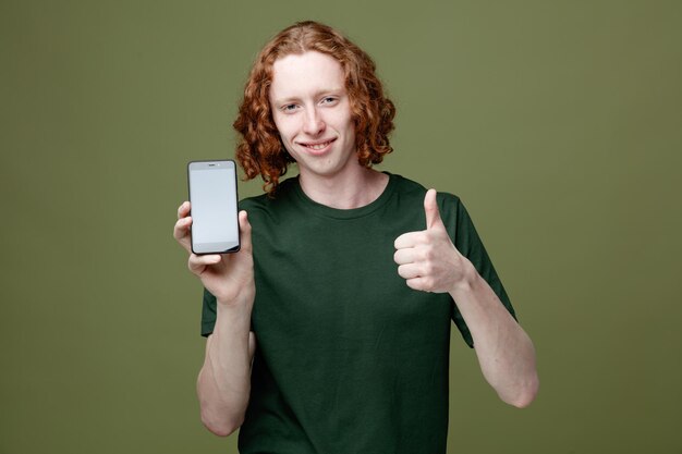 Sonriendo mostrando los pulgares hacia arriba joven guapo con camiseta verde sosteniendo el teléfono aislado sobre fondo verde