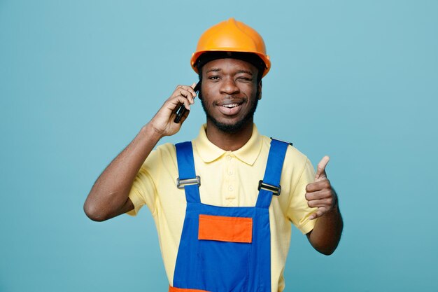 Sonriendo mostrando los pulgares hacia arriba parpadeó joven constructor afroamericano en uniforme habla por teléfono aislado sobre fondo azul.