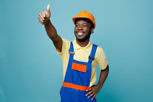 Sonriendo mostrando los pulgares hacia arriba poniendo la mano en las caderas joven constructor afroamericano en uniforme aislado sobre fondo azul.