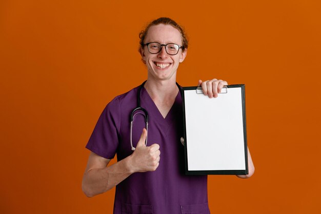 Sonriendo mostrando los pulgares hacia arriba sosteniendo portapapeles joven médico masculino vistiendo uniforme con estetoscopio aislado sobre fondo naranja