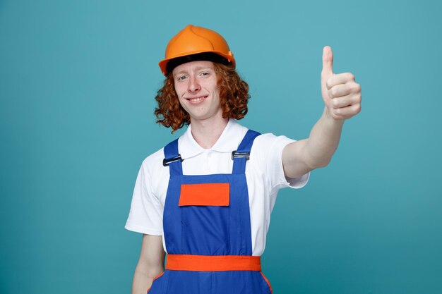 Sonriendo mostrando Thumbs up joven constructor hombre en uniforme aislado sobre fondo azul.