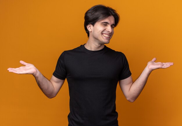 Sonriendo con los ojos cerrados chico guapo joven con camiseta negra extendiendo las manos aisladas en la pared naranja