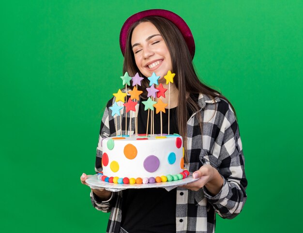 Sonriendo con los ojos cerrados hermosa joven vistiendo gorro de fiesta sosteniendo la torta