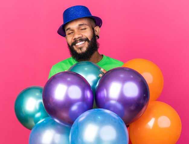 Sonriendo con los ojos cerrados joven afroamericano con sombrero de fiesta de pie detrás de globos aislados en la pared rosa