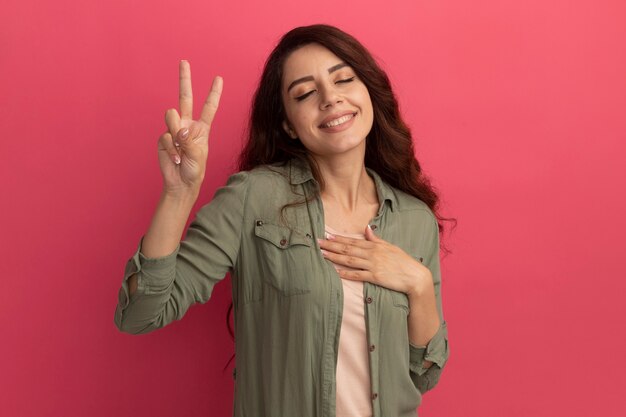 Sonriendo con los ojos cerrados joven hermosa chica con camiseta verde oliva mostrando gesto de paz aislado en la pared rosa