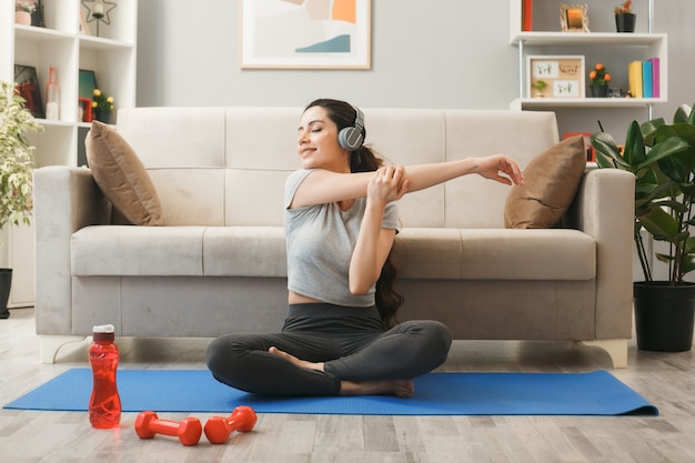 Sonriendo con los ojos cerrados niña usando audífonos haciendo ejercicio en estera de yoga delante del sofá en la sala de estar
