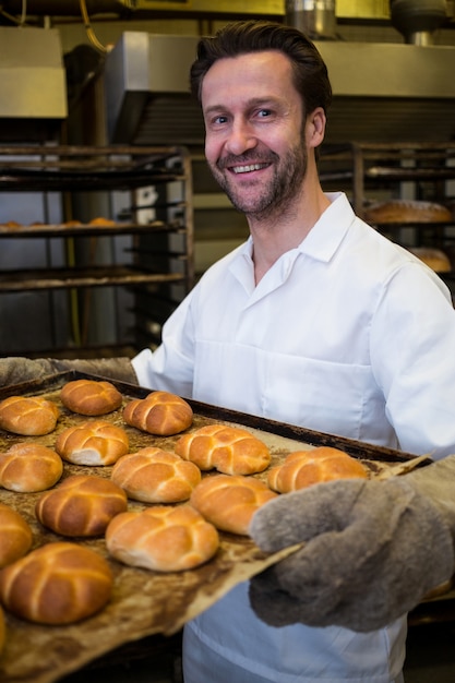 Sonriendo panadero que sostiene una bandeja de bollos recién horneados