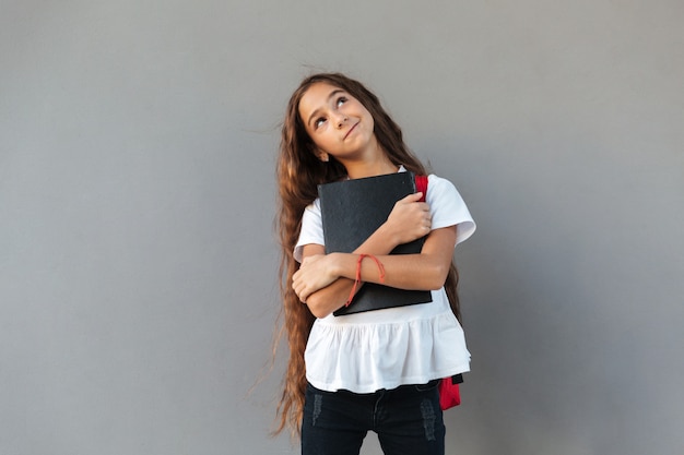 Foto gratuita sonriendo pensativa colegiala morena con libro de abrazos de pelo largo