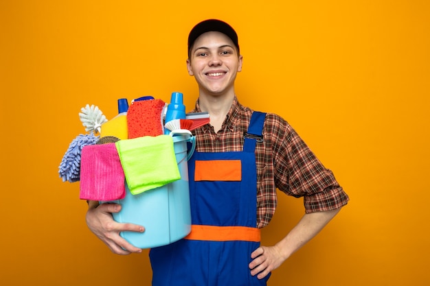 Sonriendo poniendo la mano en la cadera joven chico de limpieza con uniforme y gorra sosteniendo un balde de herramientas de limpieza