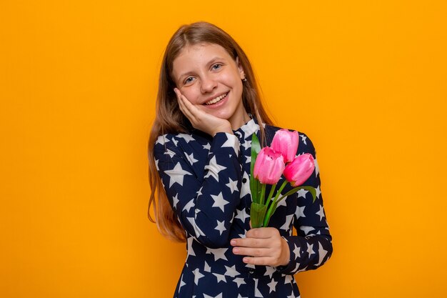 Sonriendo poniendo la mano en la mejilla hermosa niña en el día de la mujer feliz sosteniendo flores aisladas en la pared naranja