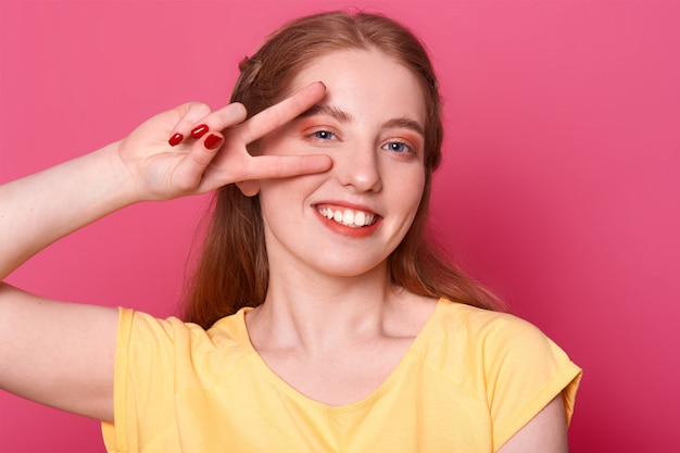 Sonriendo poses modelo positivo aislado sobre fondo rosa brillante en estudio con mano de victoria cerca de su ojo derecho, vistiendo camiseta amarilla