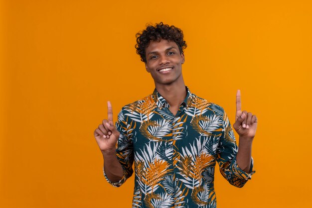 Sonriendo a positivo joven guapo de piel oscura con cabello rizado en hojas de camisa estampada apuntando con el dedo índice hacia arriba mientras mira a la cámara sobre un fondo naranja