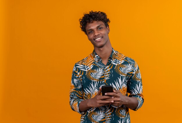 Sonriendo positivo joven guapo de piel oscura con cabello rizado en hojas camisa impresa sosteniendo el teléfono móvil mientras mira a la cámara sobre un fondo naranja