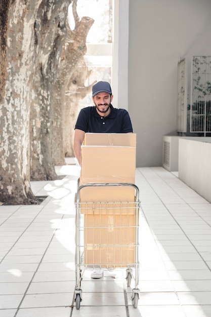 Sonriendo repartidor caminando sobre el pavimento con carro lleno de cajas de cartón