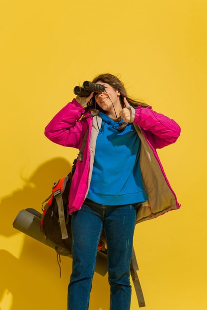 Sonriendo, se ve feliz. Retrato de una alegre joven turista caucásica con bolsa y binoculares aislado sobre fondo amarillo de estudio. Preparándose para viajar. Resort, emociones humanas, vacaciones.