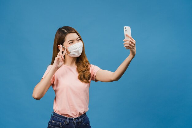 Sonriente adorable mujer asiática con mascarilla médica haciendo foto selfie en teléfono inteligente con expresión positiva en ropa casual y soporte aislado en la pared azul