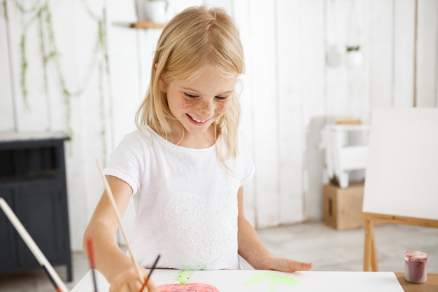 Sonriente y alegre, lleno de alegría, niño con cabello rubio y pecas sosteniendo el pincel en la mano y aspirando a pintar cuadros en la sala de arte.