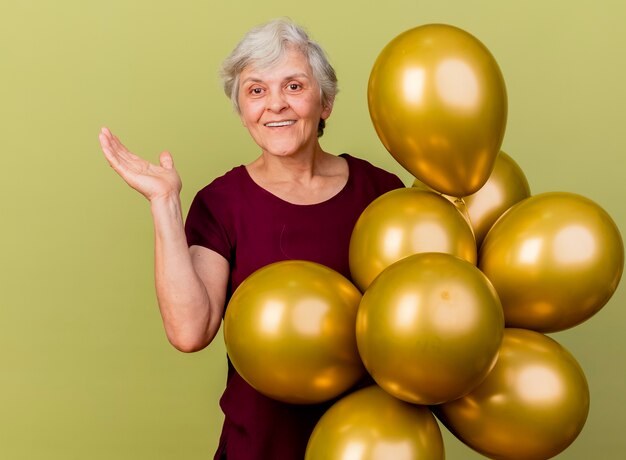 Sonriente anciana se encuentra con globos de helio sosteniendo la mano abierta aislada en la pared verde oliva con espacio de copia