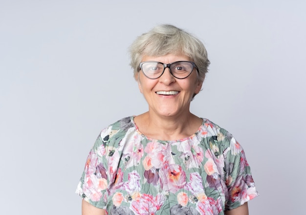 Sonriente anciana en gafas ópticas se encuentra aislado en la pared blanca