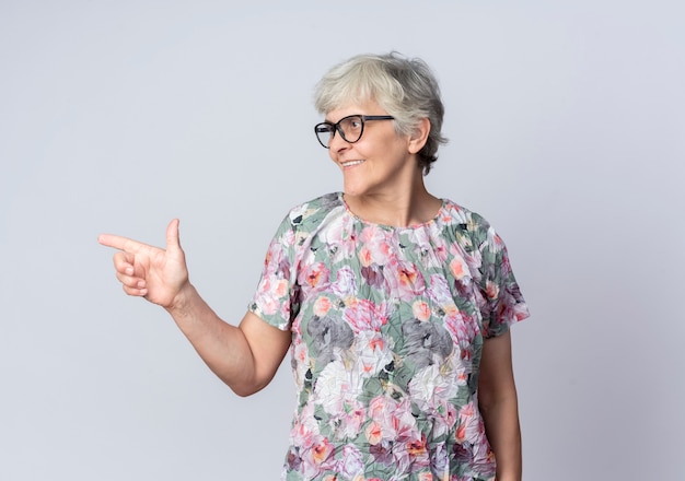 Foto gratuita sonriente anciana con gafas ópticas mira y apunta al lado aislado en la pared blanca