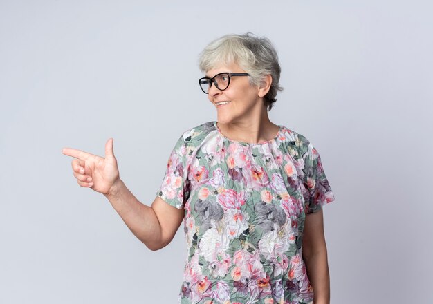 Sonriente anciana con gafas ópticas mira y apunta al lado aislado en la pared blanca